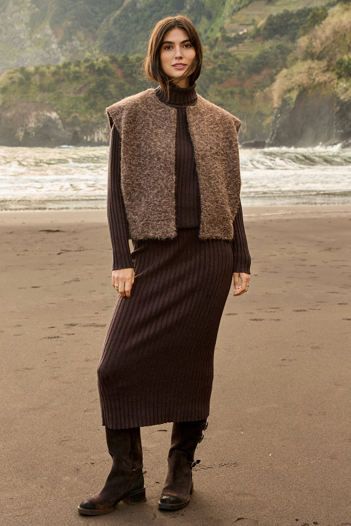 A woman stands on a beach wearing a dark brown ribbed knit midi skirt, a matching dark turtleneck, a brown faux-fur vest, and dark boots, with ocean waves and rocky cliffs in the background.