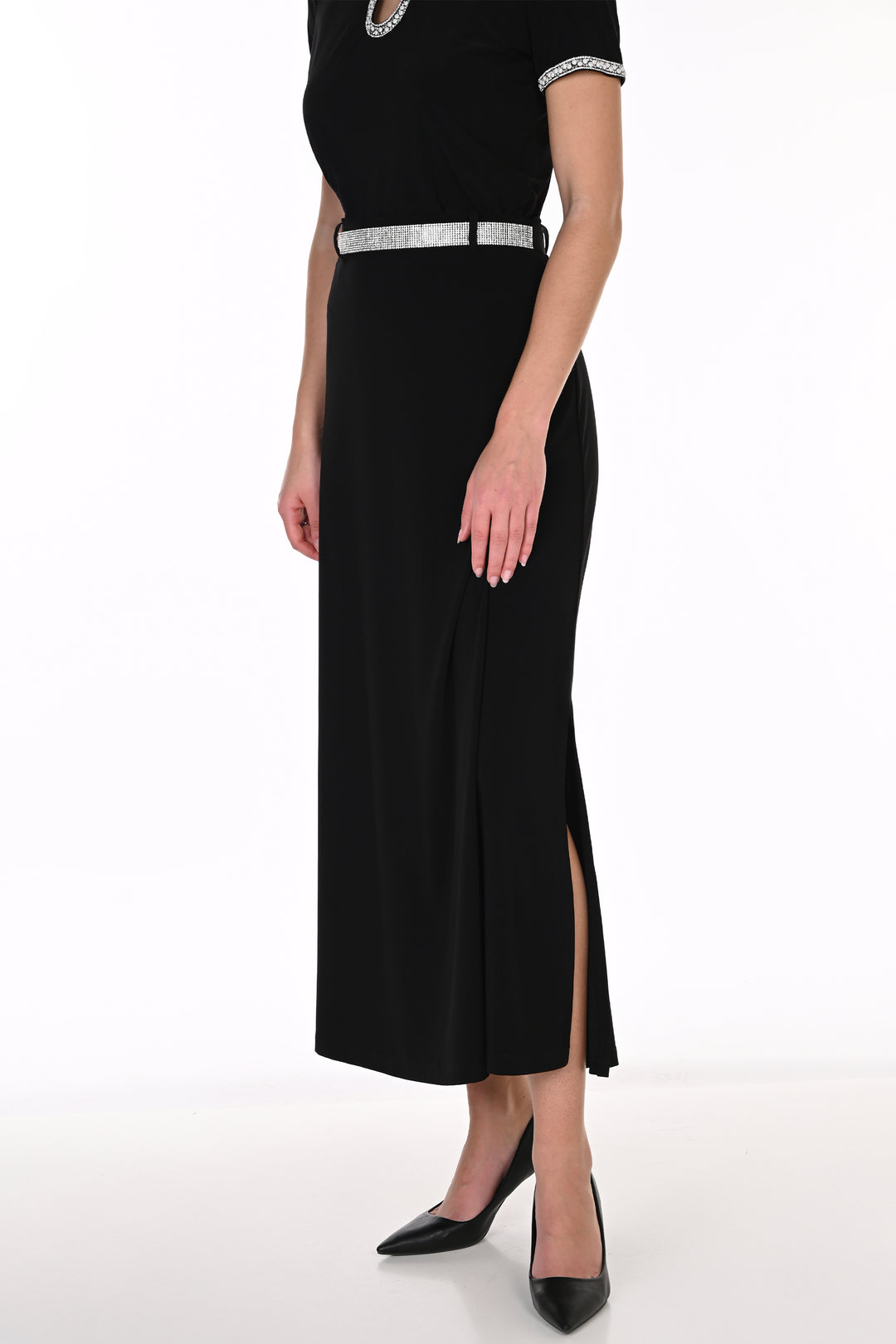 A woman modeling a black short sleeve top, midi skirt with a high slit, and a silver embellished belt, paired with black pointed-toe heels against a white studio background.