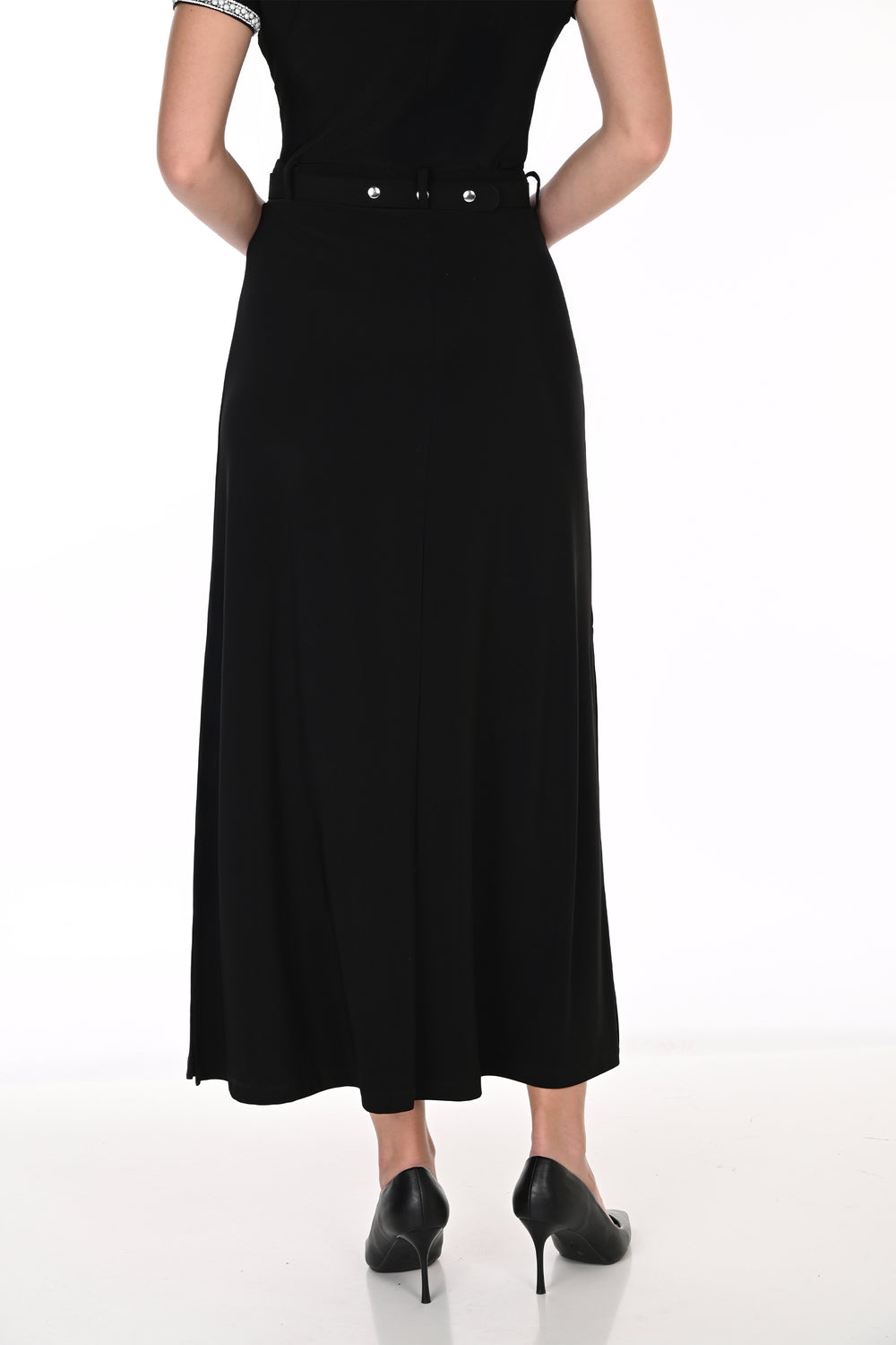 A woman modeling a black top, midi skirt with a high slit, and a silver embellished belt, paired with black pointed-toe heels against a white studio background.