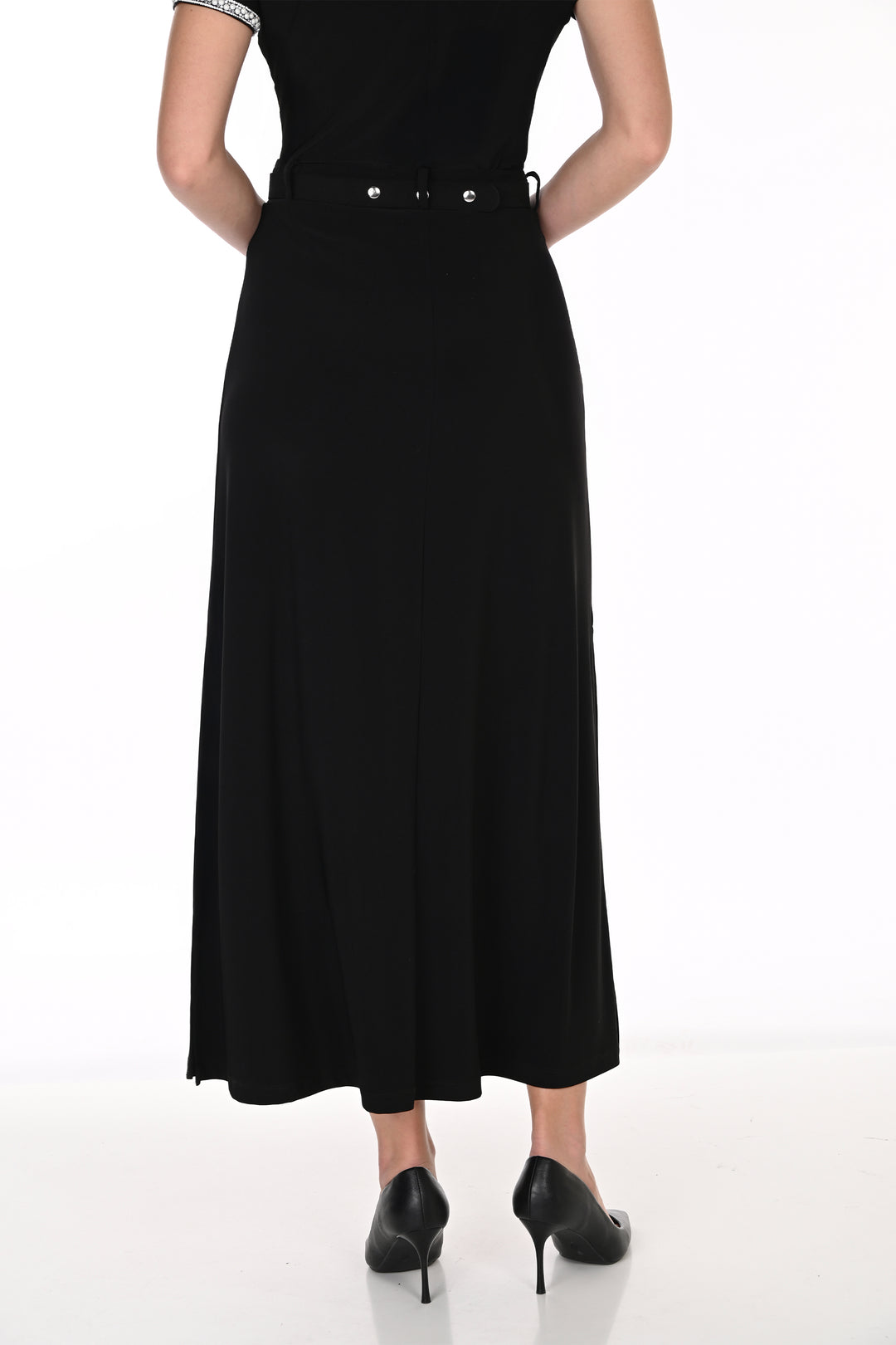 A woman modeling a black top, midi skirt with a high slit, and a silver embellished belt, paired with black pointed-toe heels against a white studio background.
