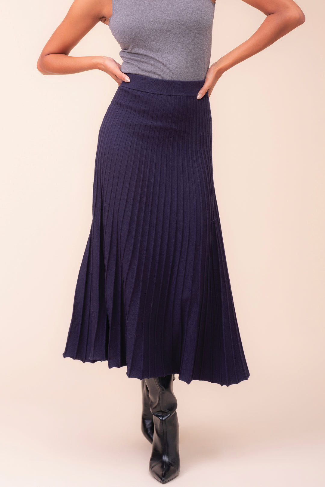 A model wearing a dark navy blue pleated midi skirt, paired with a gray sleeveless top and black pointed-toe ankle boots, standing against a solid light beige background.