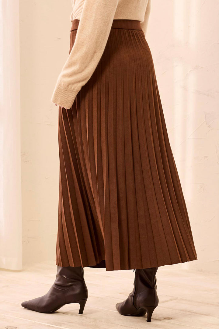A woman modelling a brown pleated suede skirt and beige sweater in front of white background. 