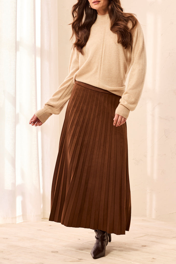 A woman modelling a brown pleated suede skirt and beige sweater in front of white background. 