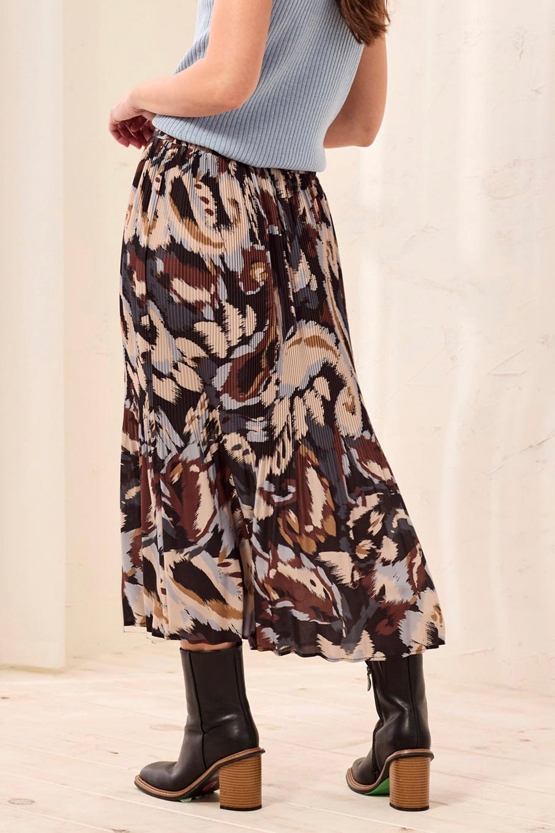 A woman shown from the back wearing a brown and beige printed textured shirt and black boots standing in front of a beige background. 