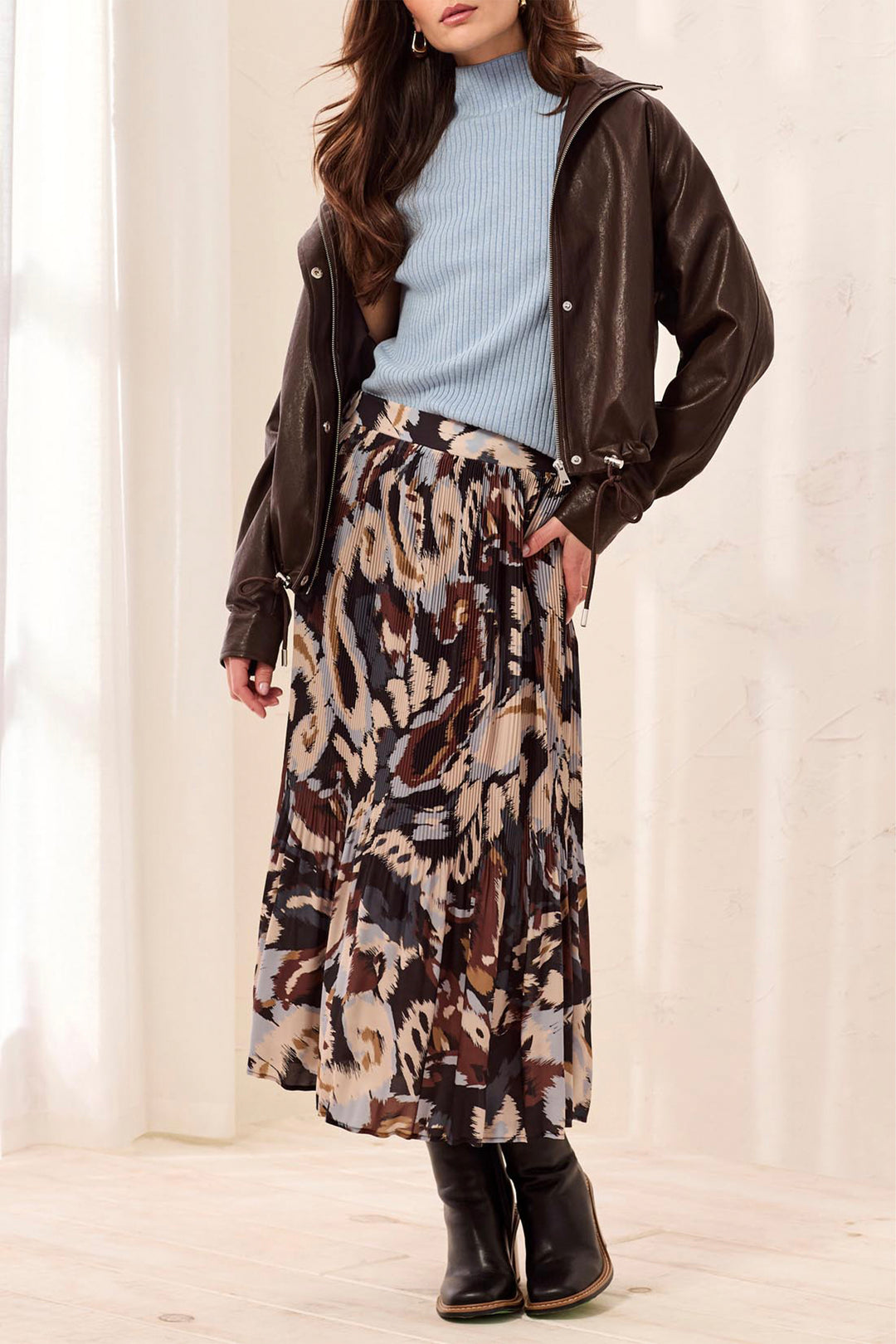 A woman wearing a brown and beige printed textured shirt and black boots standing in front of a beige background. 