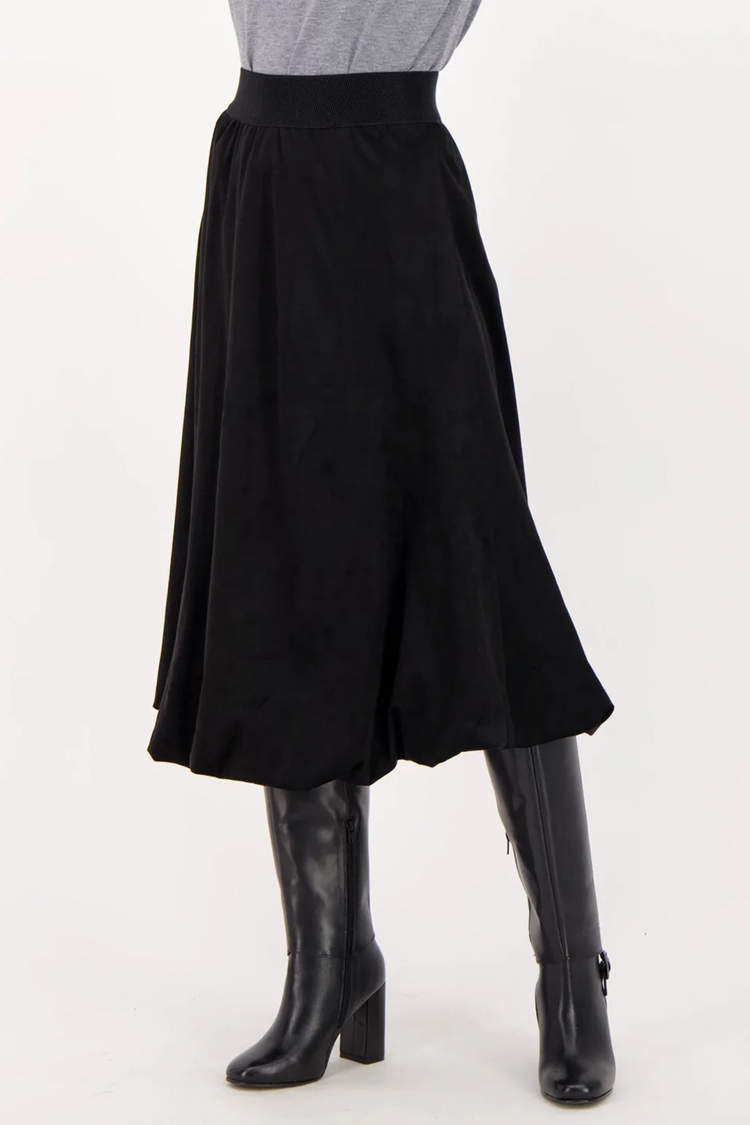 A woman wearing a long black suede skirt over black boots standing in front of a grey background. 