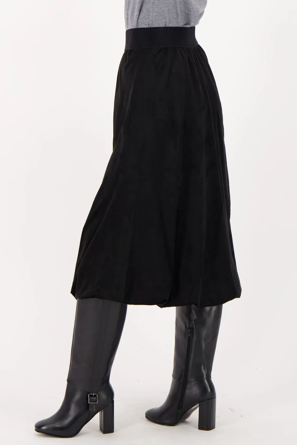 A woman shown from the back wearing a long black suede skirt over black boots standing in front of a grey background. 