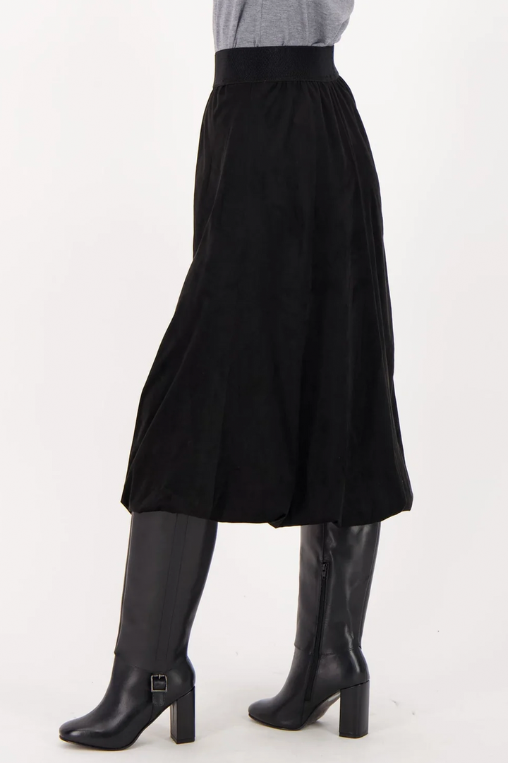 A woman shown from the back wearing a long black suede skirt over black boots standing in front of a grey background. 