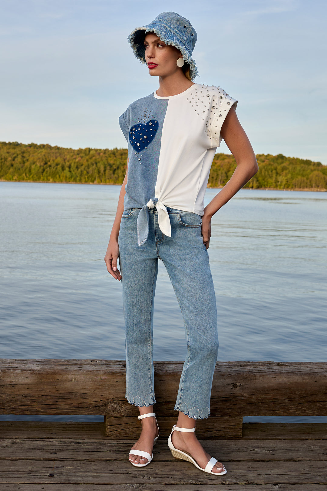 Woman standing on a dock by a lake wearing a blue and white outfit with a denim hat.