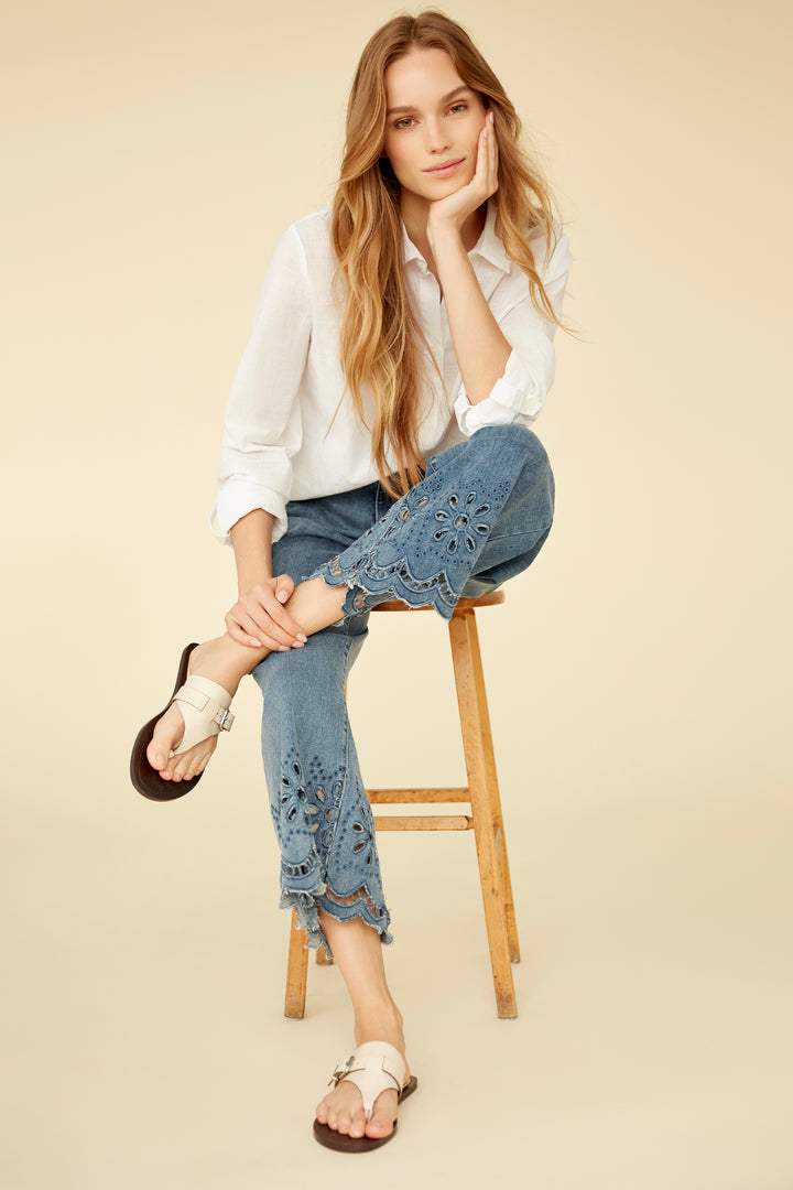 Woman sitting on a wooden stool wearing a white blouse and blue jeans with lace details.