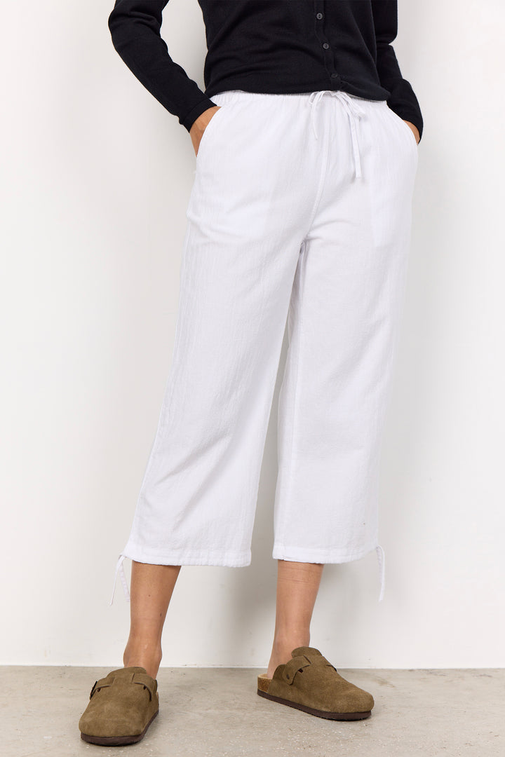 Person wearing white wide-leg crop pants with tie hems and brown shoes on a plain background