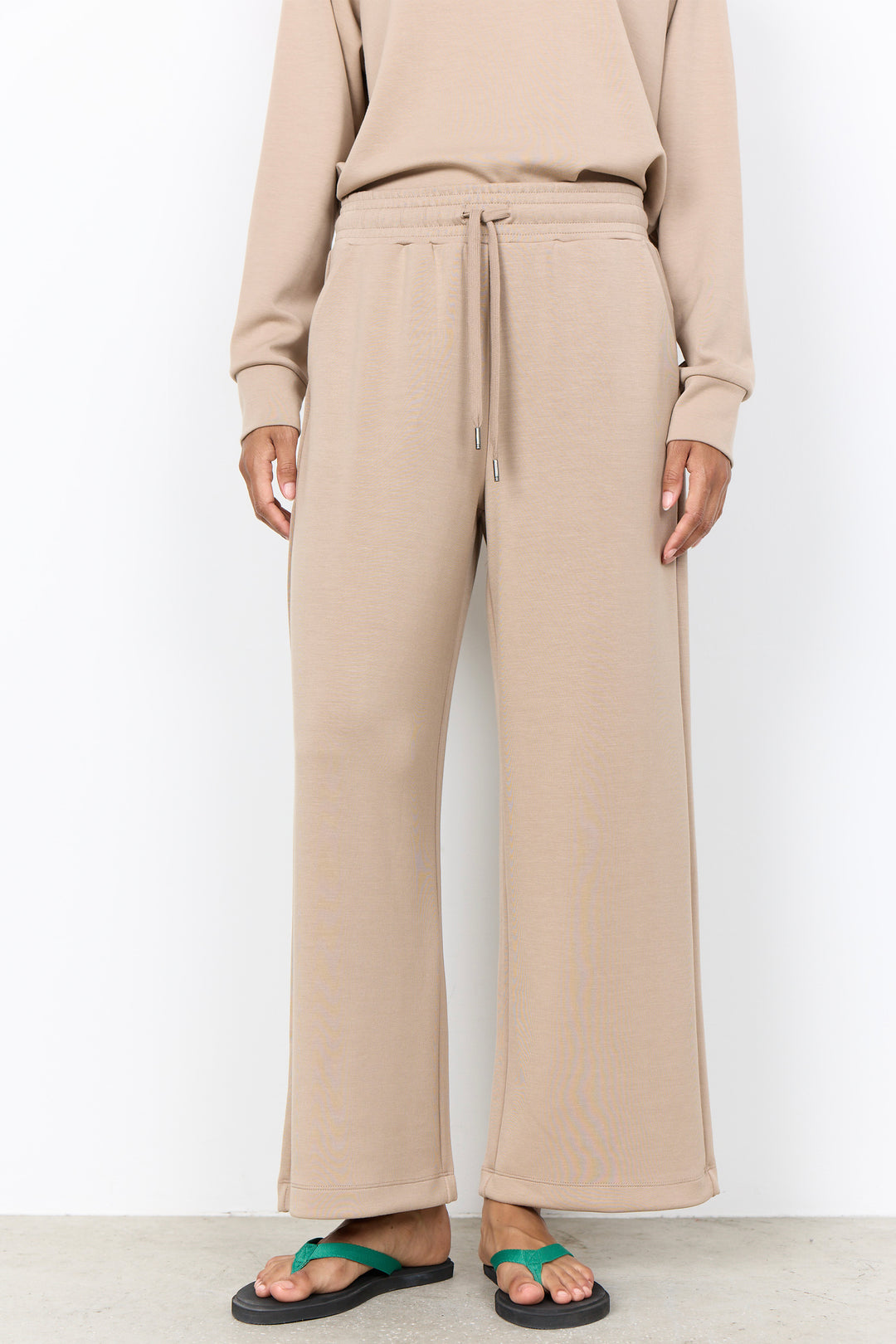 A person wearing beige wide-leg sweatpants with a drawstring waist, paired with a matching beige sweatshirt and green flip-flop sandals, shown against a plain white studio background.