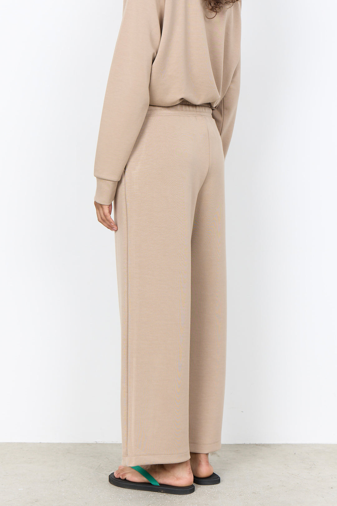 A person shown from the back wearing a matching beige long-sleeve top and wide-leg pants set, with black sandals, against a plain light gray studio background.