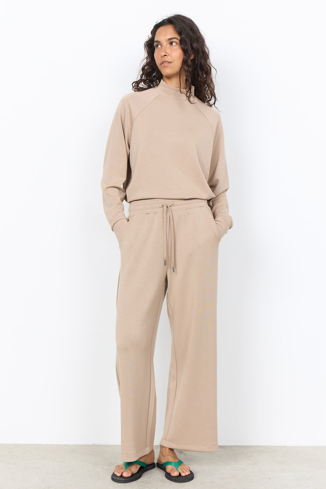 A woman modeling a matching beige lounge set consisting of a long-sleeve mock-neck top and wide-leg drawstring pants, paired with green sandals against a white studio background.
