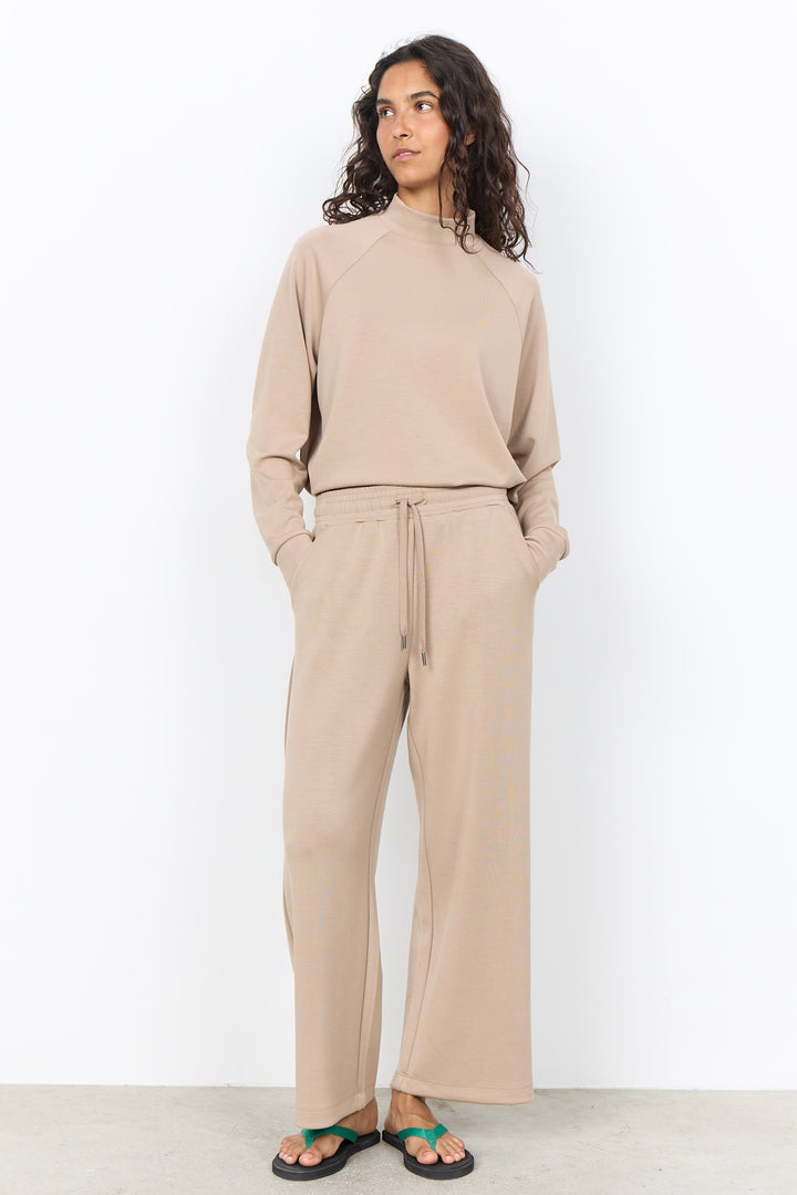 A woman modeling a matching beige lounge set consisting of a long-sleeve mock-neck top and wide-leg drawstring pants, paired with green sandals against a white studio background.