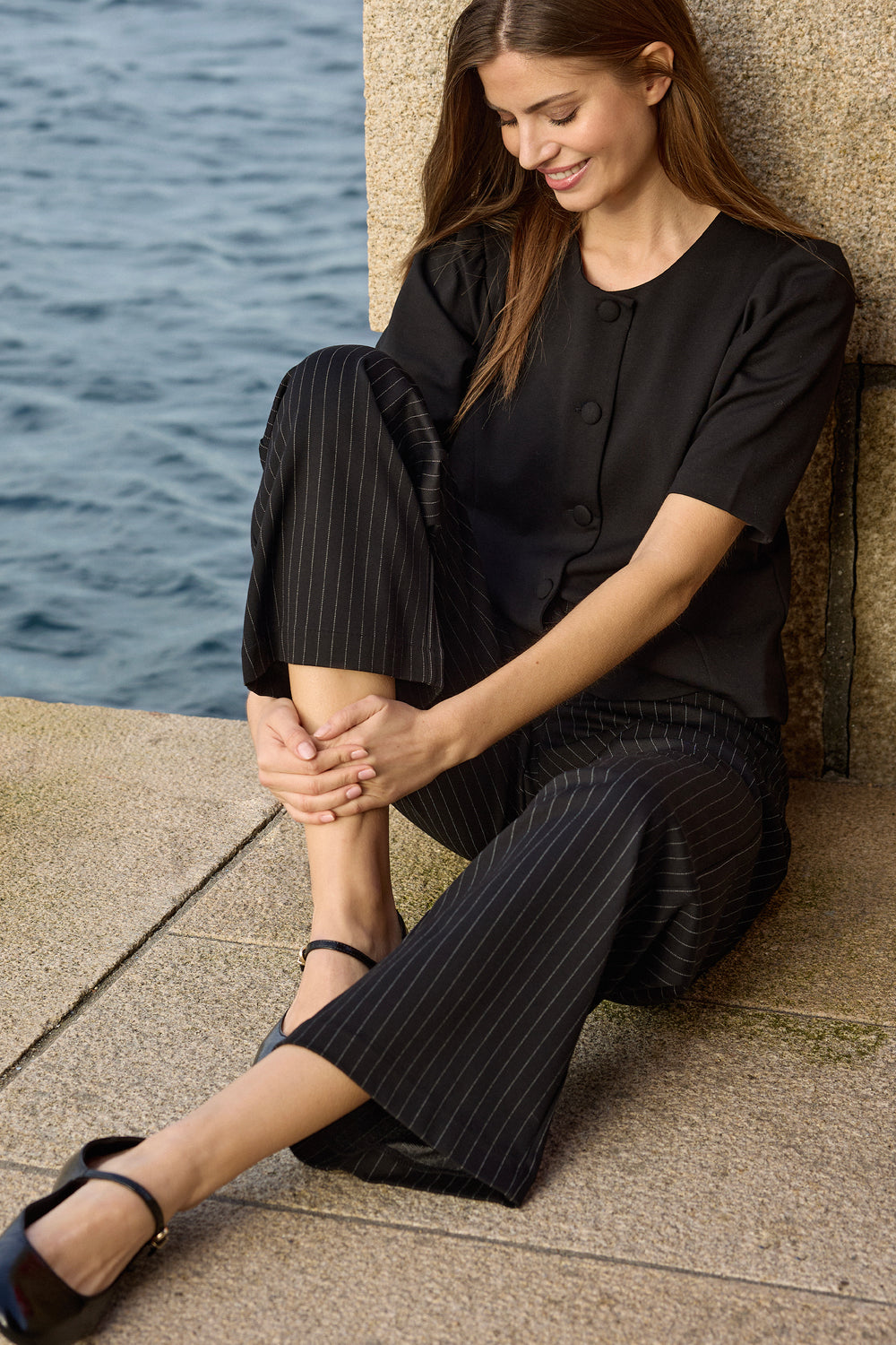 woman wearing a black blouse and black pinstripe wide leg pants sitting by water.