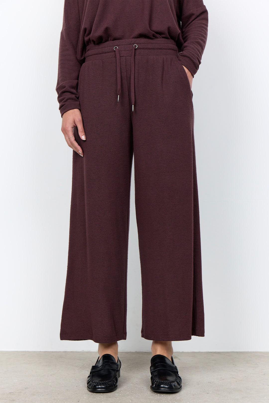 A person wearing loose, wide-leg cropped pants in a deep plum-burgundy color, paired with a matching long-sleeve top and black loafers, standing against a plain light gray wall.