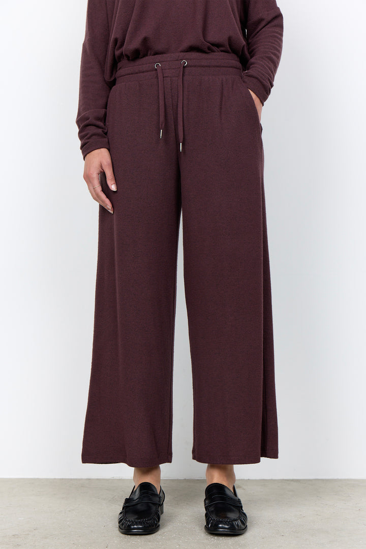A person wearing loose, wide-leg cropped pants in a deep plum-burgundy color, paired with a matching long-sleeve top and black loafers, standing against a plain light gray wall.