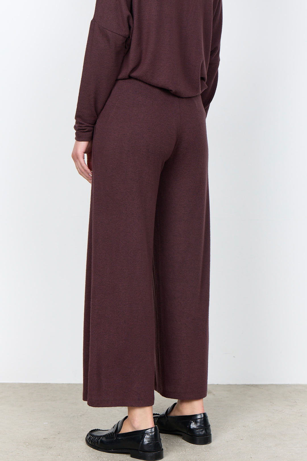 A person shown from the back wearing a matching dark plum-brown long-sleeve top and wide-leg cropped pants, paired with black loafers, against a plain light gray wall and concrete floor.