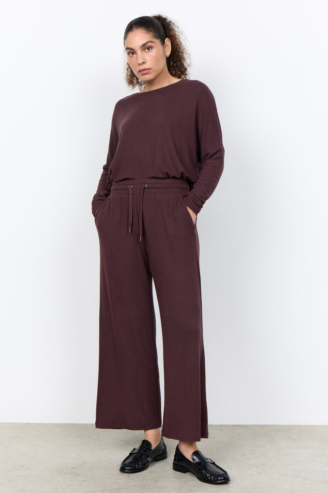 A woman modeling a matching two-piece lounge set in a deep plum-burgundy color, featuring a long-sleeve top and wide-leg cropped pants with a drawstring waist, paired with black loafers against a clean white studio background.