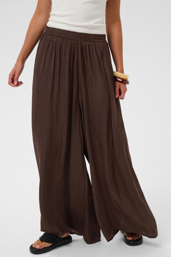 A person wearing loose, wide-leg brown pants with a gathered elastic waistband, paired with a white top and black sandals, photographed against a light gray studio background.