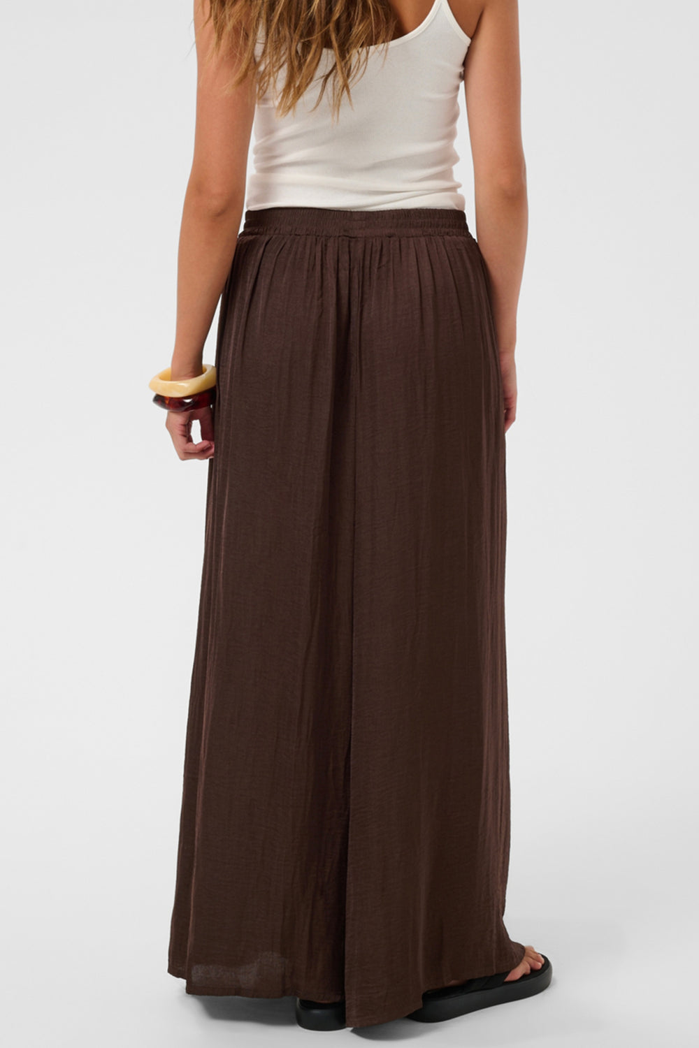 A woman shown from the back wearing a long, flowing dark brown maxi skirt and a white tank top against a light gray studio background.