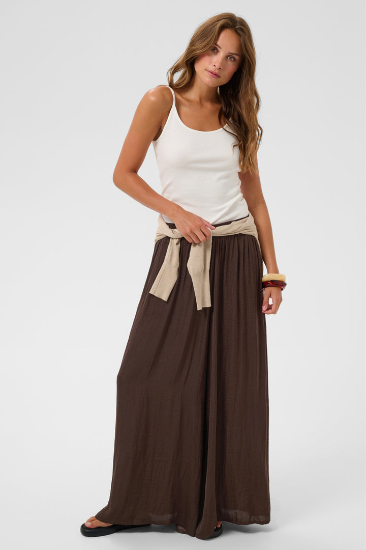 A woman modeling a white ribbed tank top and a long brown maxi skirt with a beige tie belt, standing against a light gray studio background.