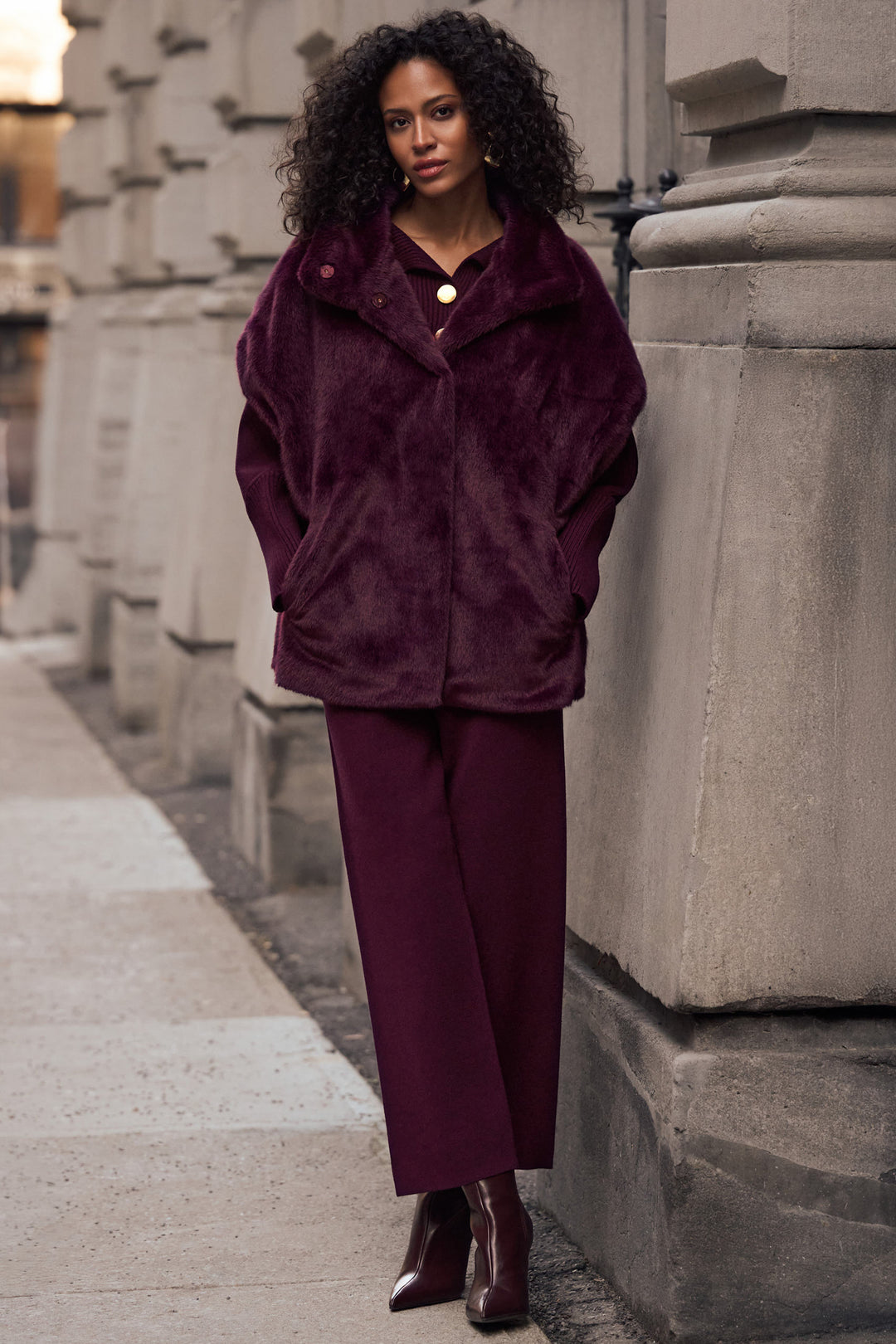 A woman stands on a city sidewalk beside a stone building, wearing a plush deep burgundy faux-fur jacket with a collar and front buttons, matching wide-leg trousers, and dark brown ankle boots.