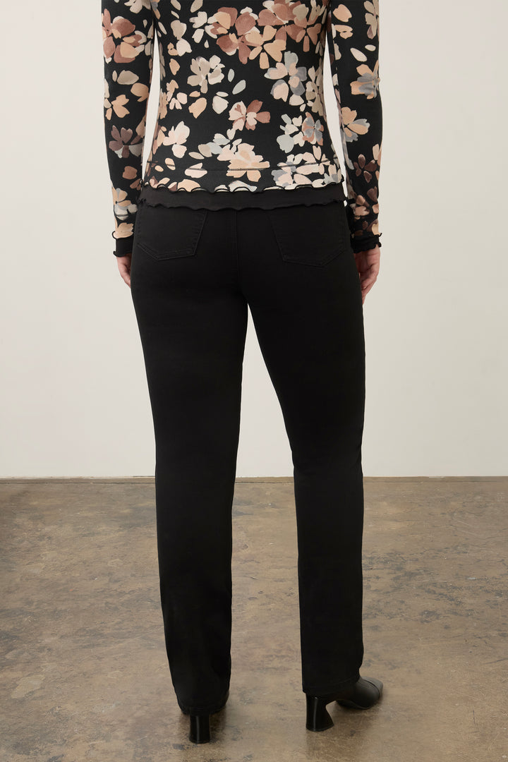 Rear view of a model wearing black trousers and a black floral long-sleeve top, standing against a plain off-white wall and concrete floor.
