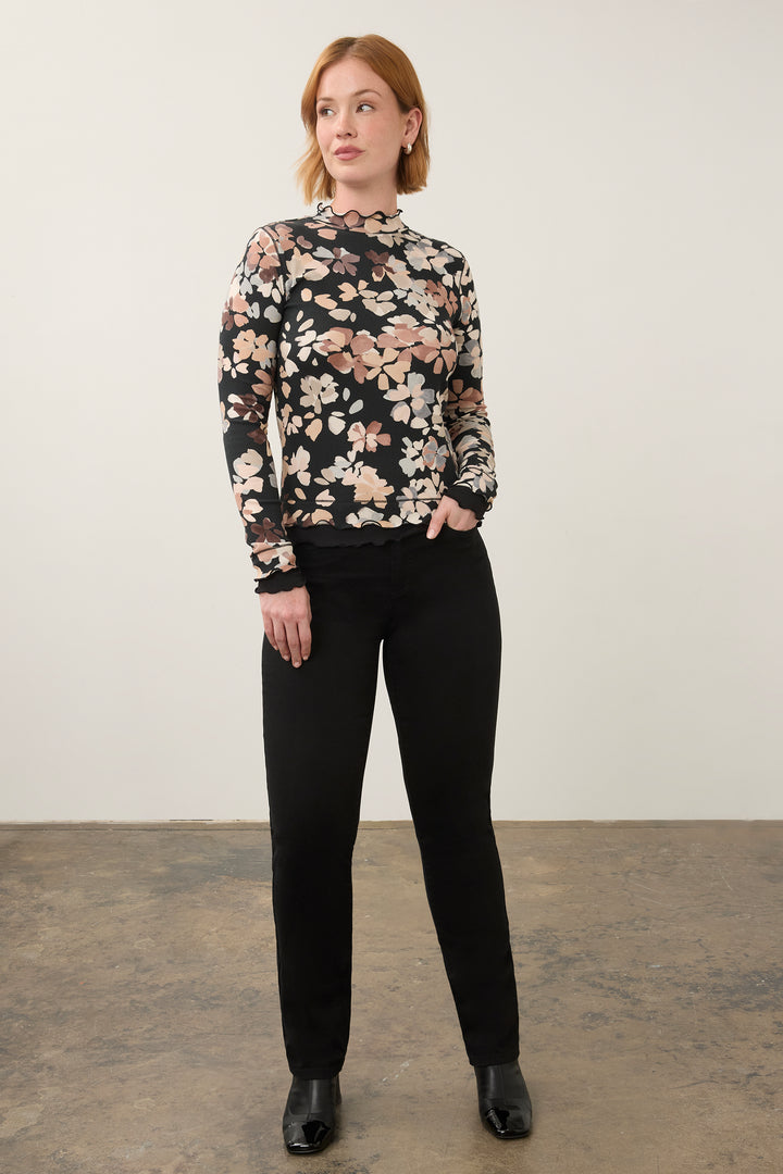 A woman modeling a black long-sleeve blouse with a beige and pink floral pattern, paired with black trousers and black shoes, standing against a plain off-white wall.