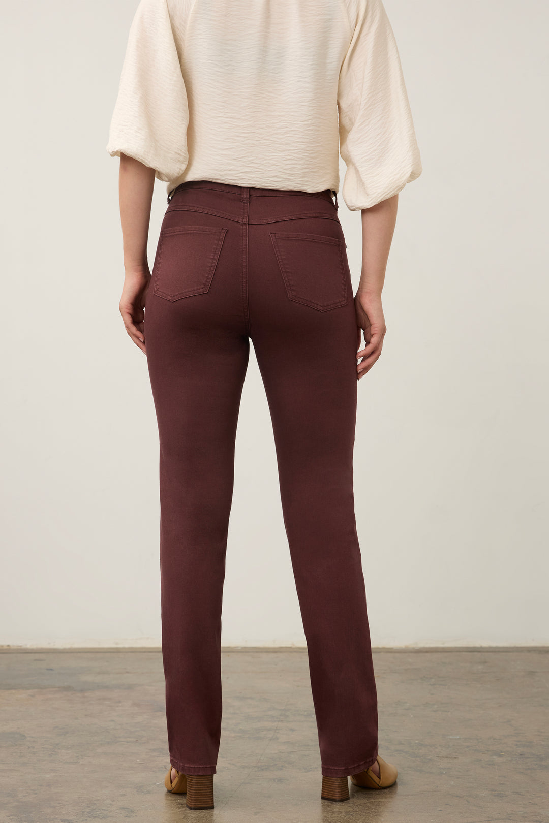 A person shown from the back wearing slim-fit brownish-burgundy pants, a cream puff-sleeve top, and brown shoes against a plain light wall and concrete floor.