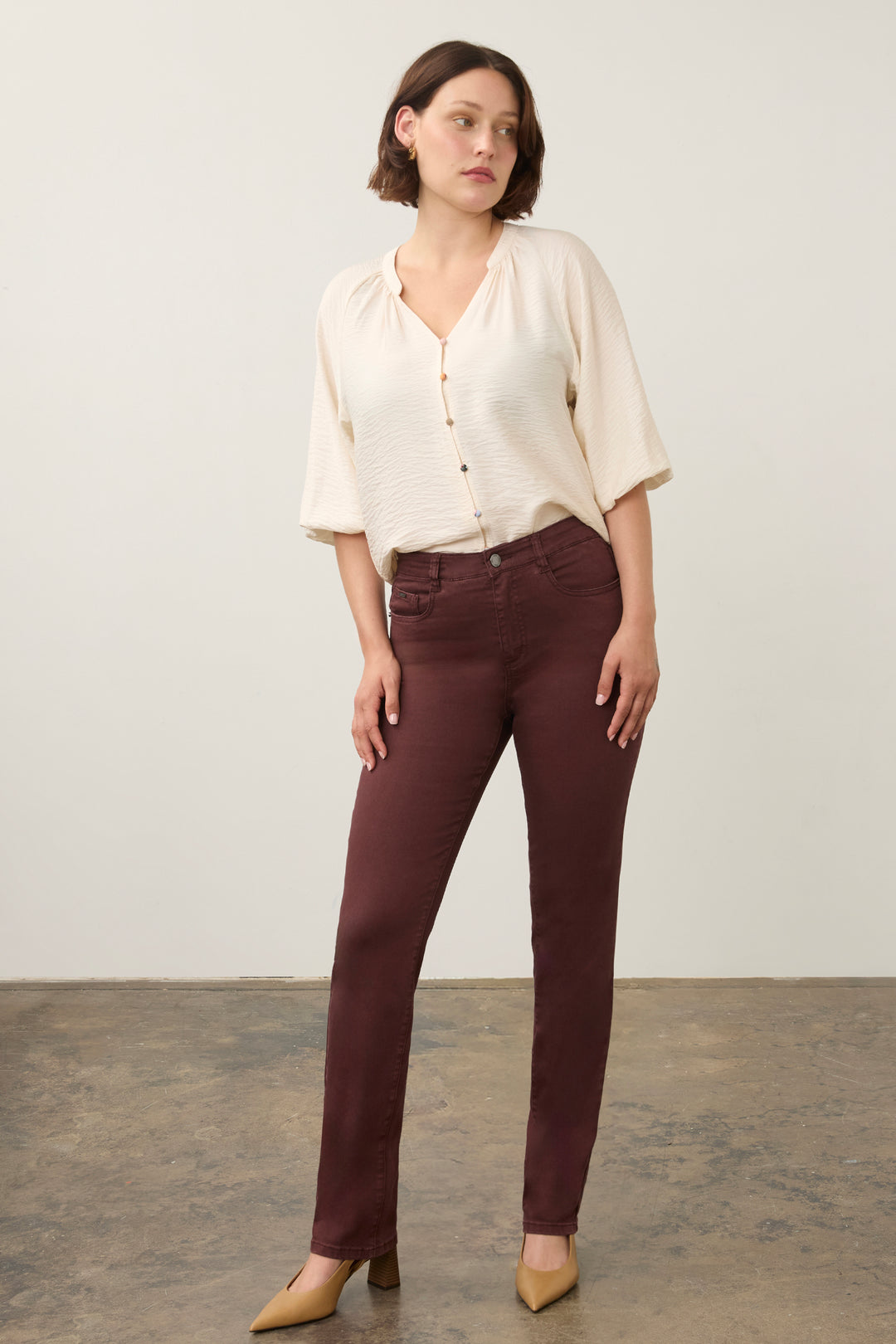 A woman modeling a cream-colored short-sleeve button-up shirt and dark brown slim-fit trousers, paired with beige heels, standing against a neutral studio backdrop.