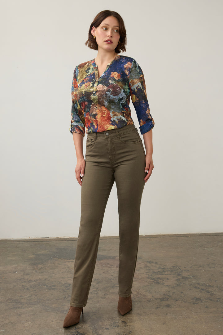 A woman modeling a three-quarter sleeve blouse with a vibrant multicolor abstract print, paired with olive green trousers and brown pointed-toe heels, standing against a plain off-white wall.