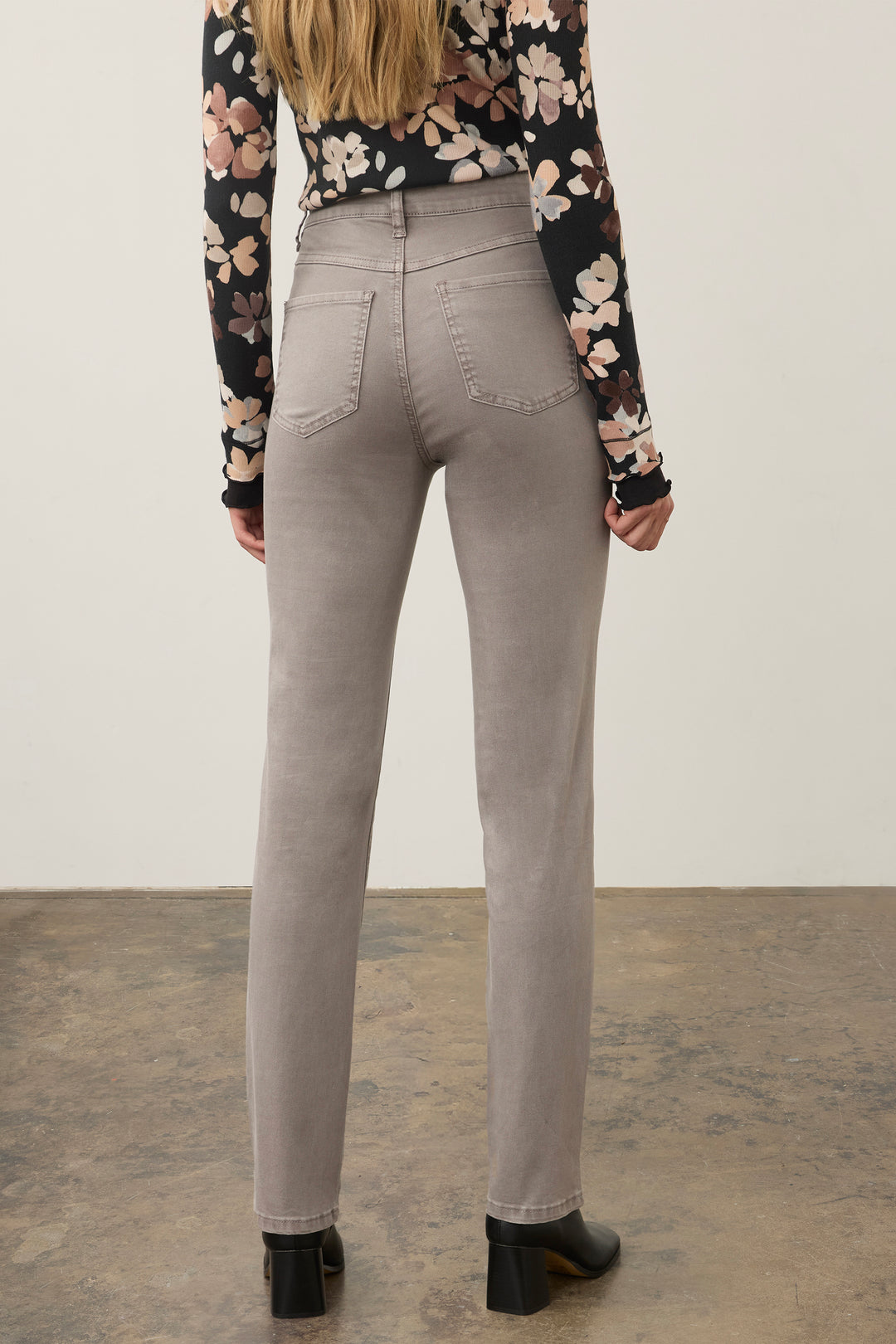 A model shown from the back wearing light gray slim-fit jeans, a black floral long-sleeve top, and black boots, standing against a plain off-white wall and concrete floor.