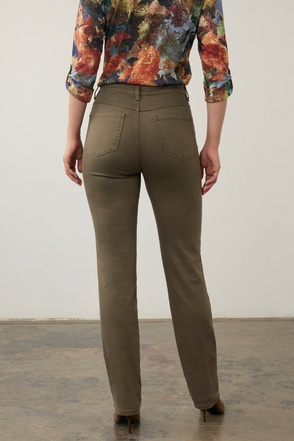 Rear view of a person wearing olive green straight-leg pants and a colorful floral long-sleeve blouse, standing against a plain light wall.