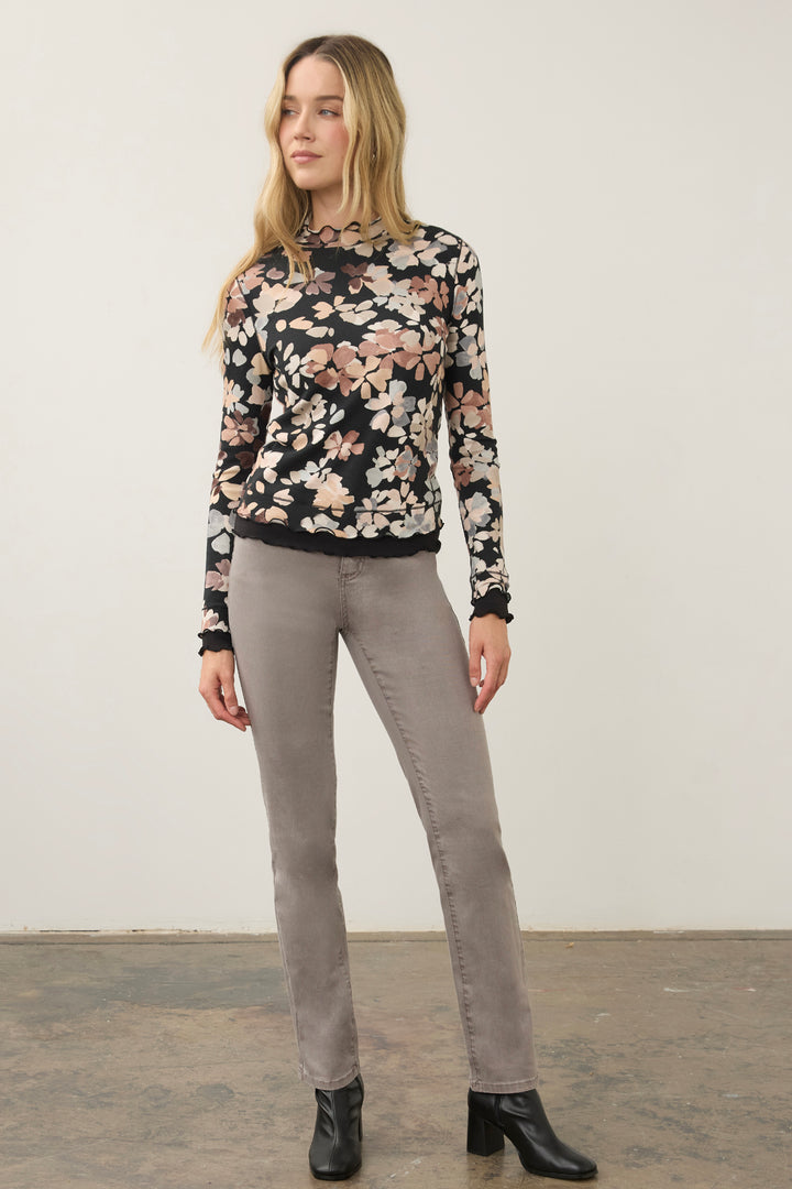 A woman modeling a black long-sleeve floral mock-neck top with beige and pink flowers, paired with gray pants and black ankle boots, standing against a plain off-white wall.