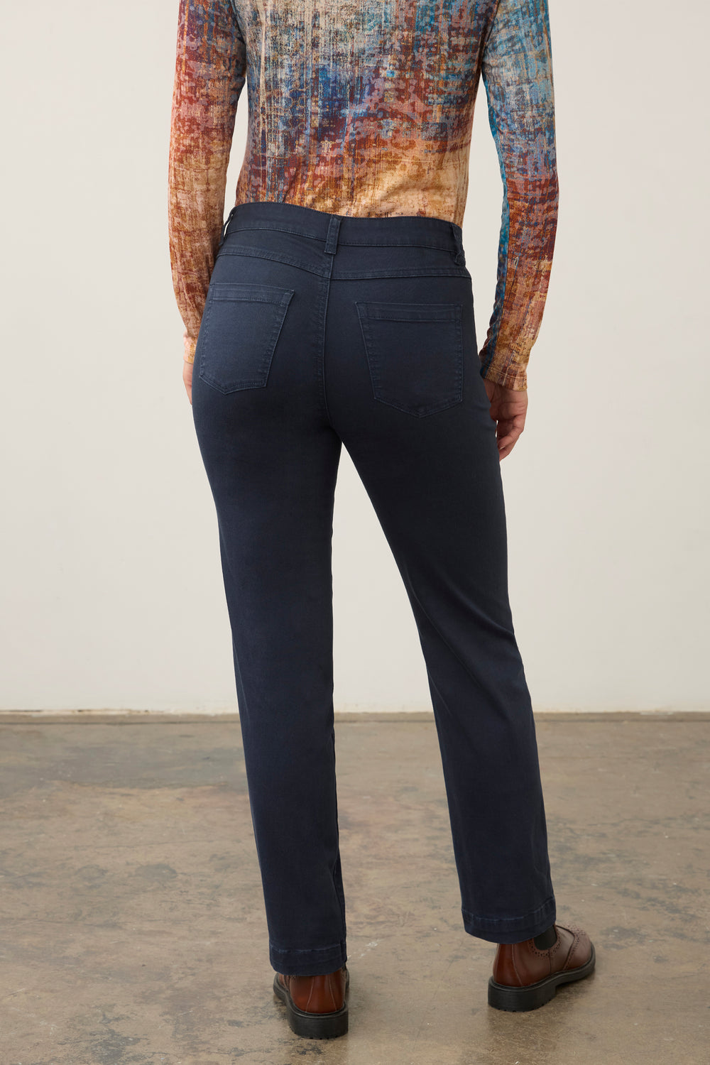 A person shown from the back wearing dark navy pants, a multicolored patterned long-sleeve top, and brown shoes against a plain indoor wall.