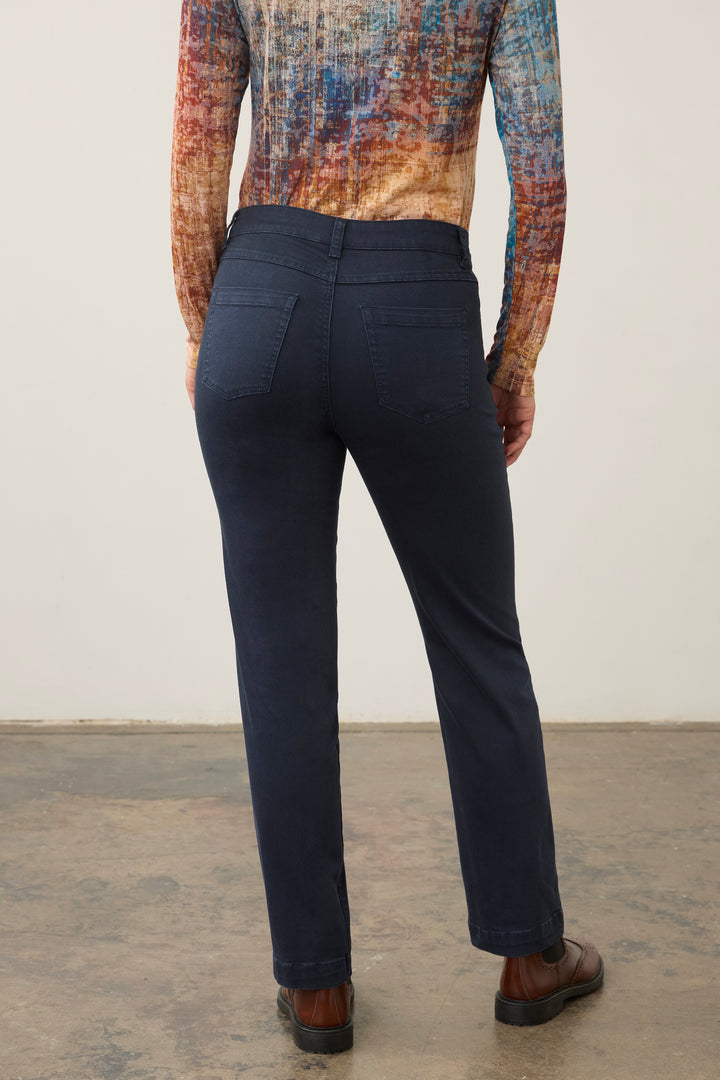 A person shown from the back wearing dark navy pants, a multicolored patterned long-sleeve top, and brown shoes against a plain indoor wall.