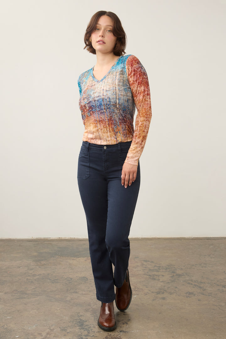 A woman modeling a long-sleeve V-neck knit top with a multicolor abstract pattern in blue, orange, and beige, paired with dark blue jeans and brown leather shoes, standing against a plain light gray wall.