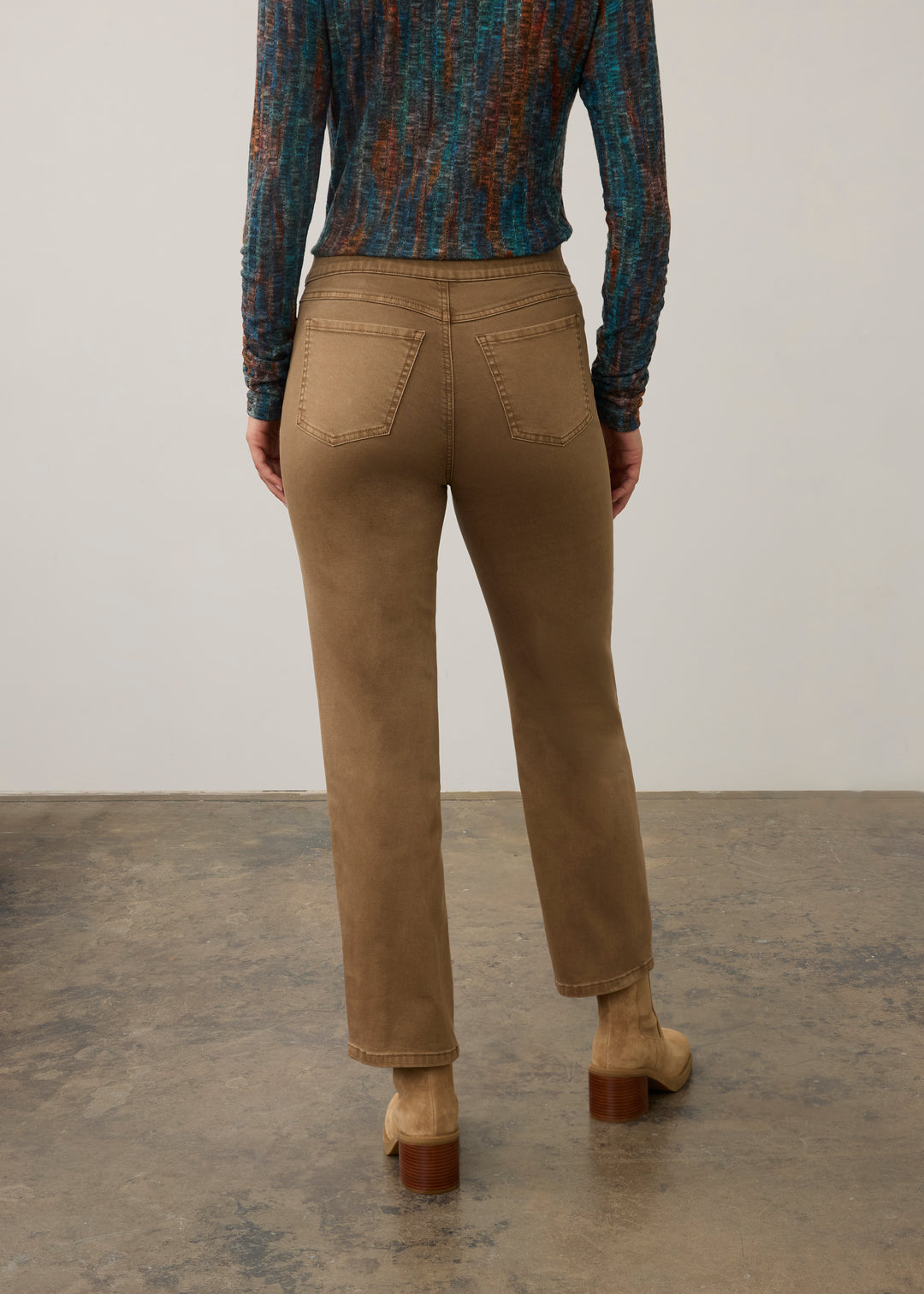 A person shown from the back wearing straight-leg brown pants, a patterned long-sleeve top, and brown heeled ankle boots against a plain studio wall and concrete floor.
