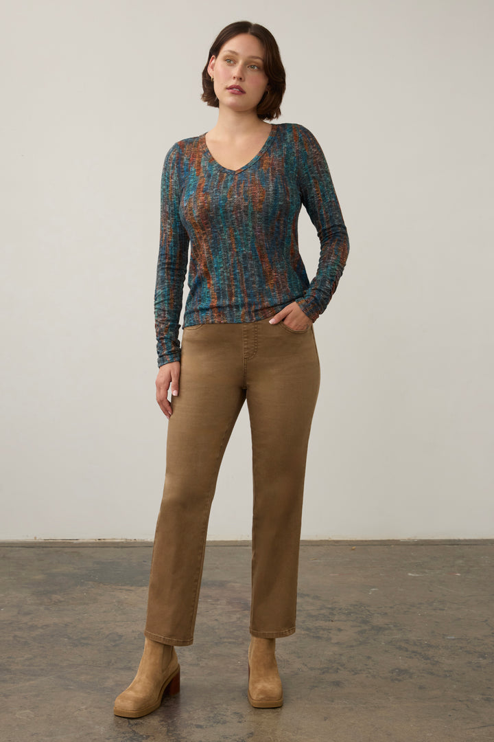A woman modeling a long-sleeve V-neck knit top with an abstract blue, teal, and rust pattern, paired with brown pants and tan ankle boots, standing against a plain light gray wall.