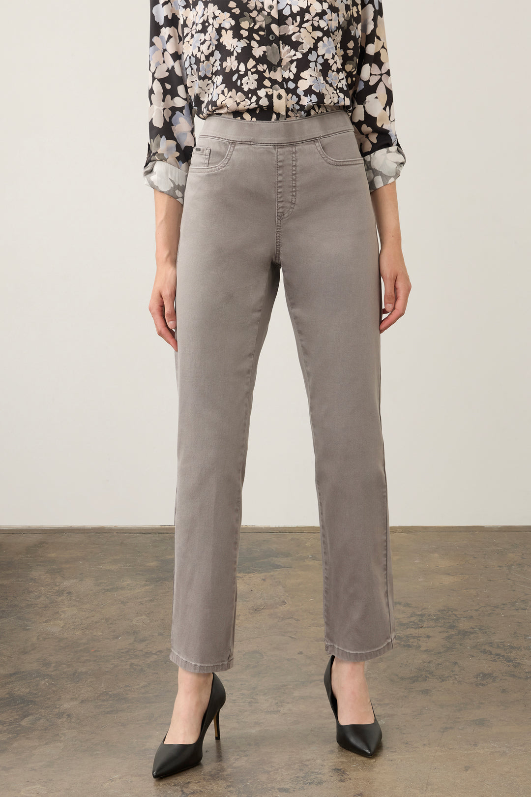 A model is shown from the waist down wearing high-waisted, straight-leg gray trousers, a black and white floral blouse, and black pointed-toe heels against a neutral studio backdrop.
