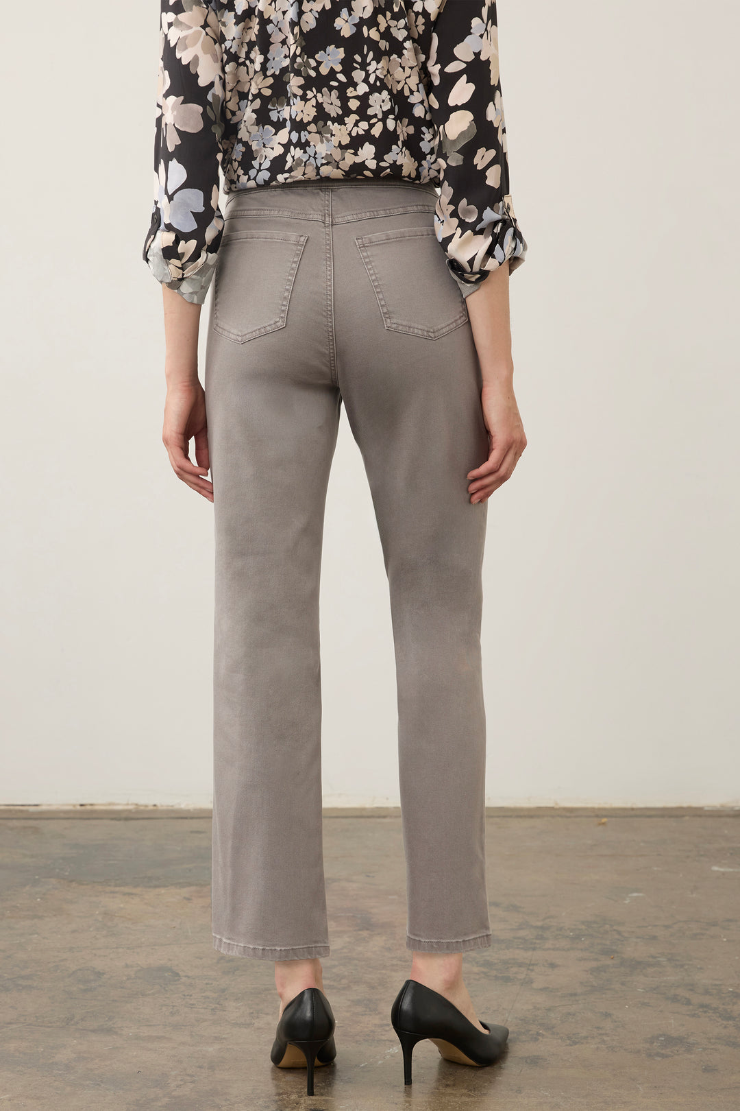 A person shown from the back wearing light gray cropped trousers, a black floral blouse, and black heels against a plain off-white wall and concrete floor.