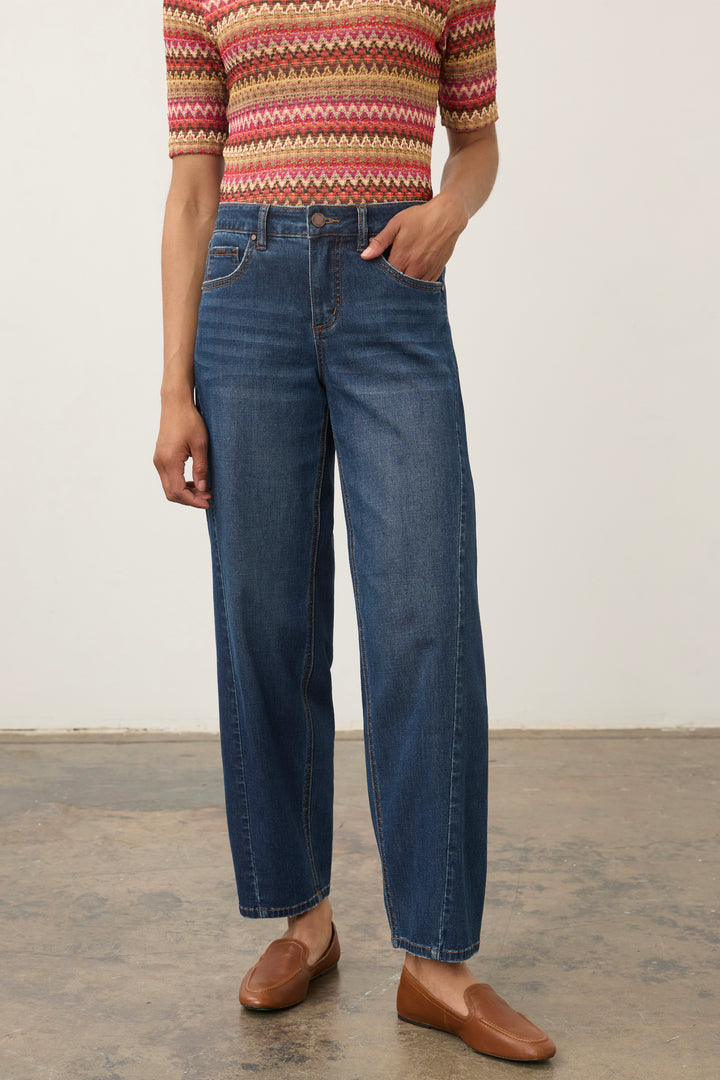 A person wearing blue straight-leg jeans, a short-sleeve knit top with a red, beige, and brown geometric pattern, and brown leather loafers, standing against a plain off-white wall and concrete floor.
