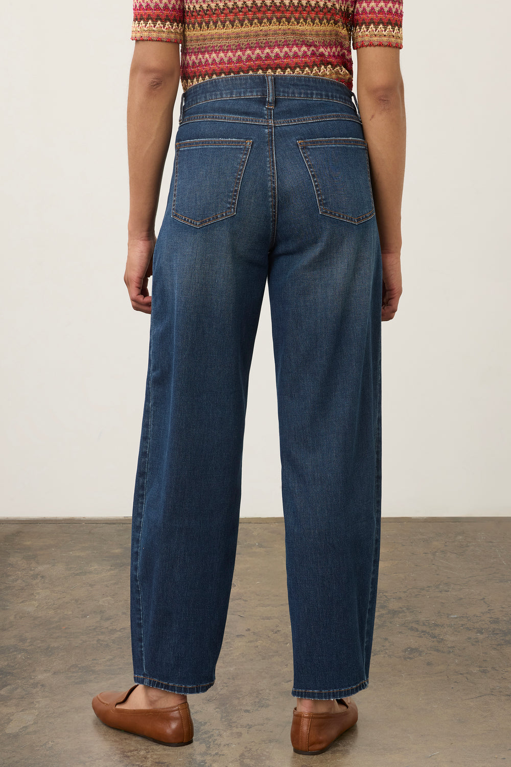 Rear view of a person wearing dark blue wide-leg jeans, a patterned short-sleeve top, and brown leather loafers against a plain light wall and concrete floor.