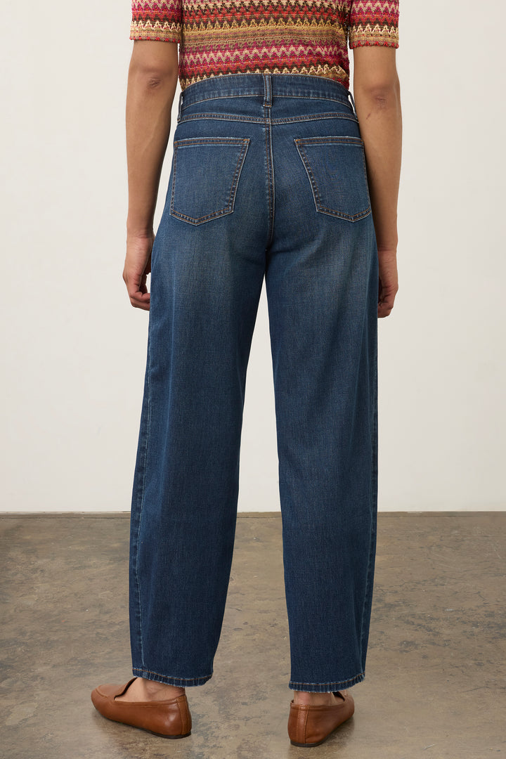 Rear view of a person wearing dark blue wide-leg jeans, a patterned short-sleeve top, and brown leather loafers against a plain light wall and concrete floor.