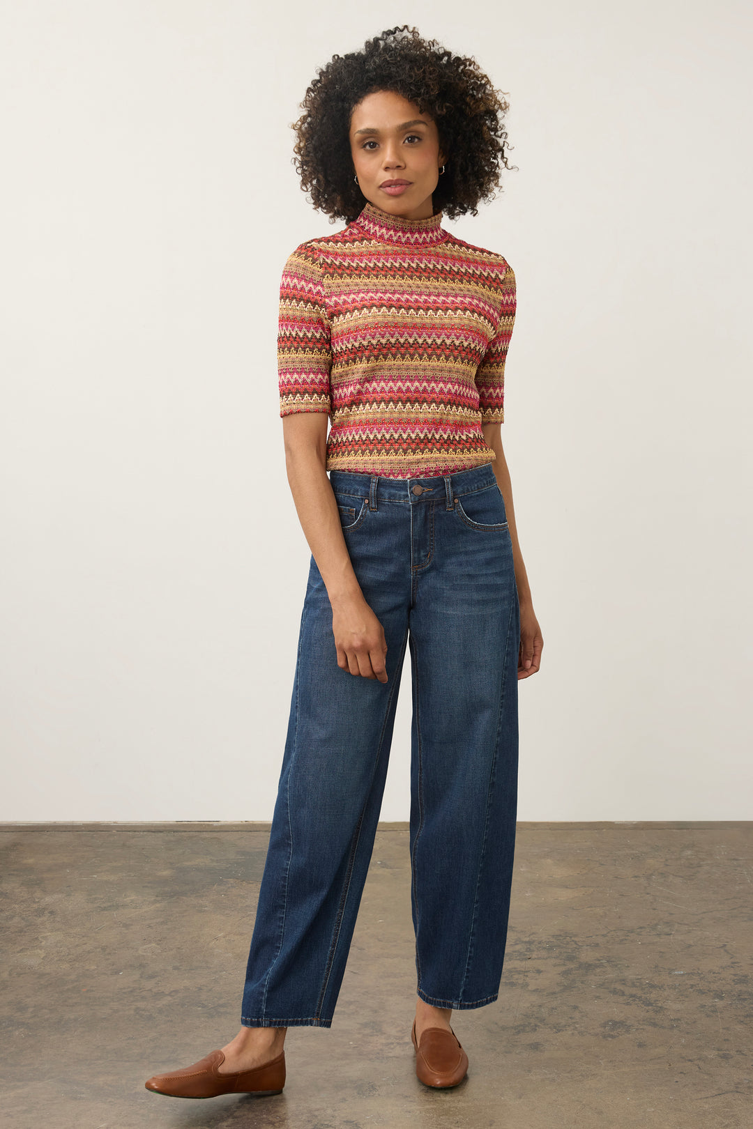 A woman modeling a short-sleeve mock-neck knit top with a multicolor geometric pattern in red, pink, yellow, and white, paired with dark blue wide-leg jeans and brown slip-on shoes, against a plain studio background.