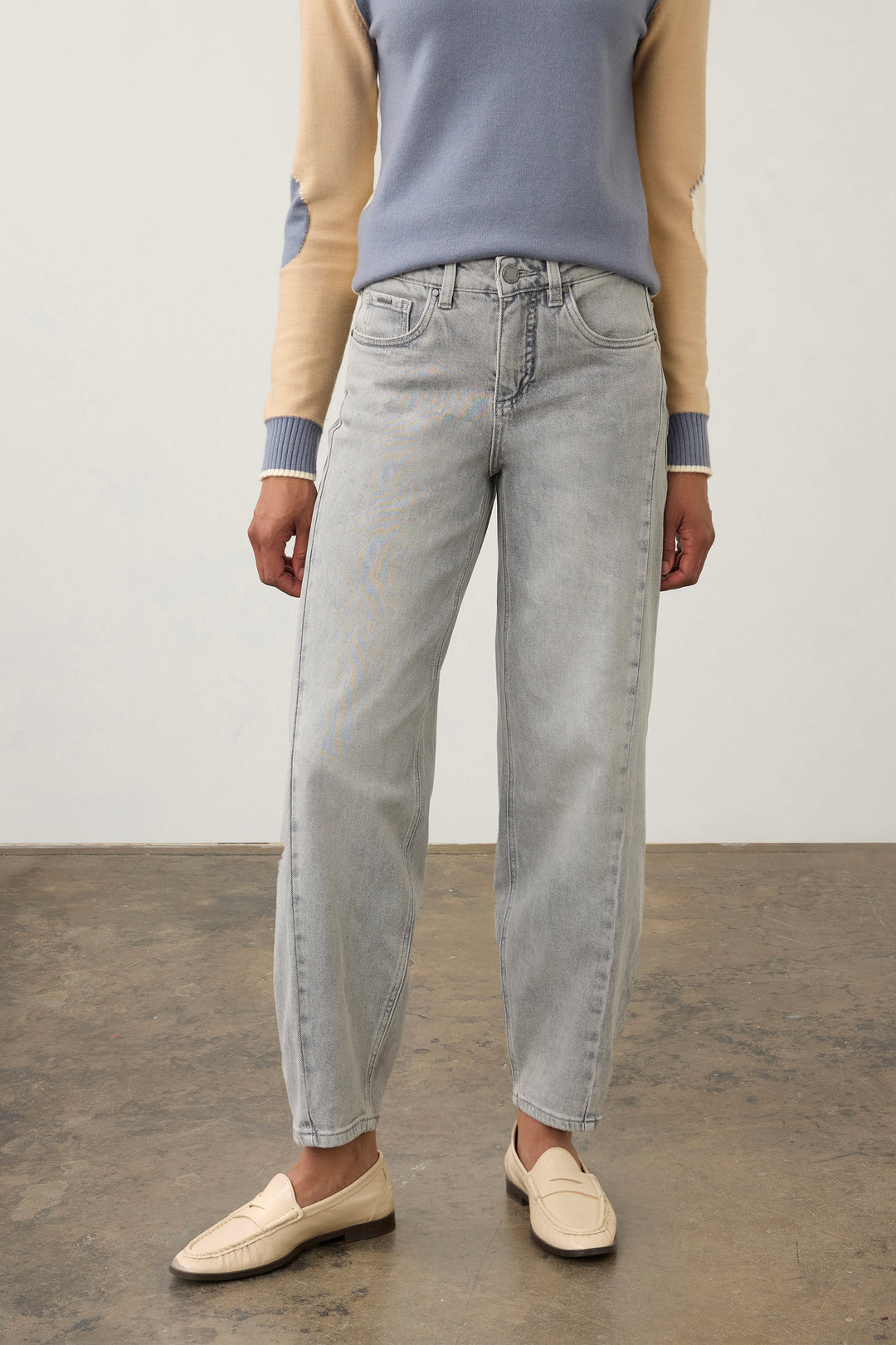A person wearing light-wash, straight-leg denim jeans with a beige and blue knit sweater and cream loafers, standing against a plain studio wall.