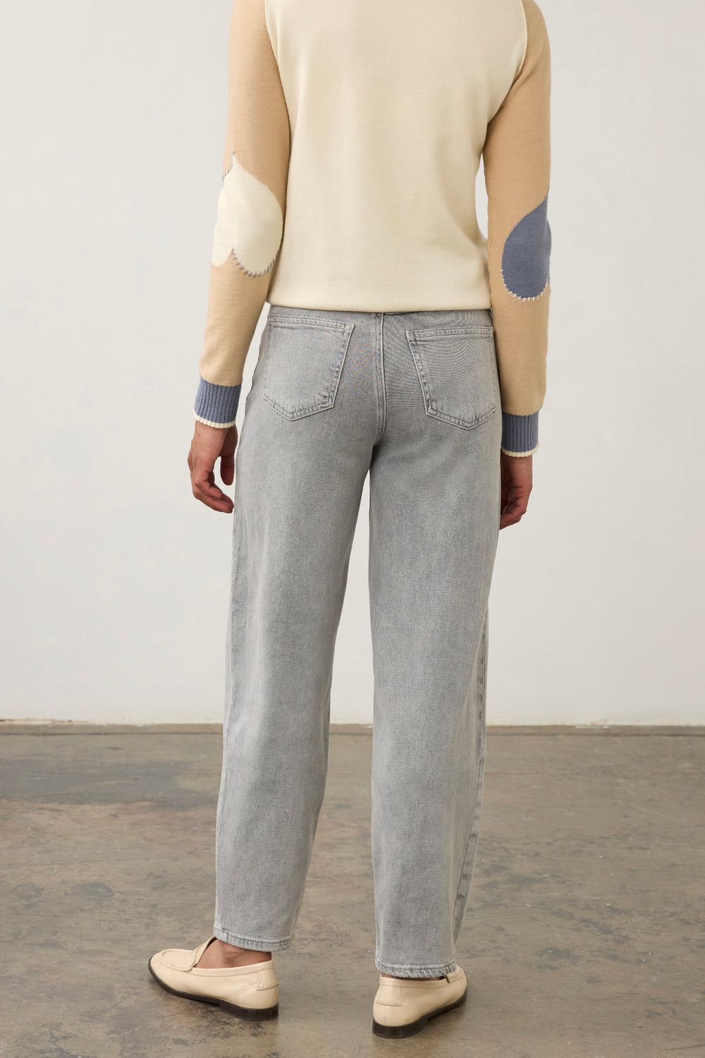 A person shown from the back wearing light gray straight-leg jeans, a cream sweater with beige and blue patch details, and cream loafers, standing against a plain light wall.