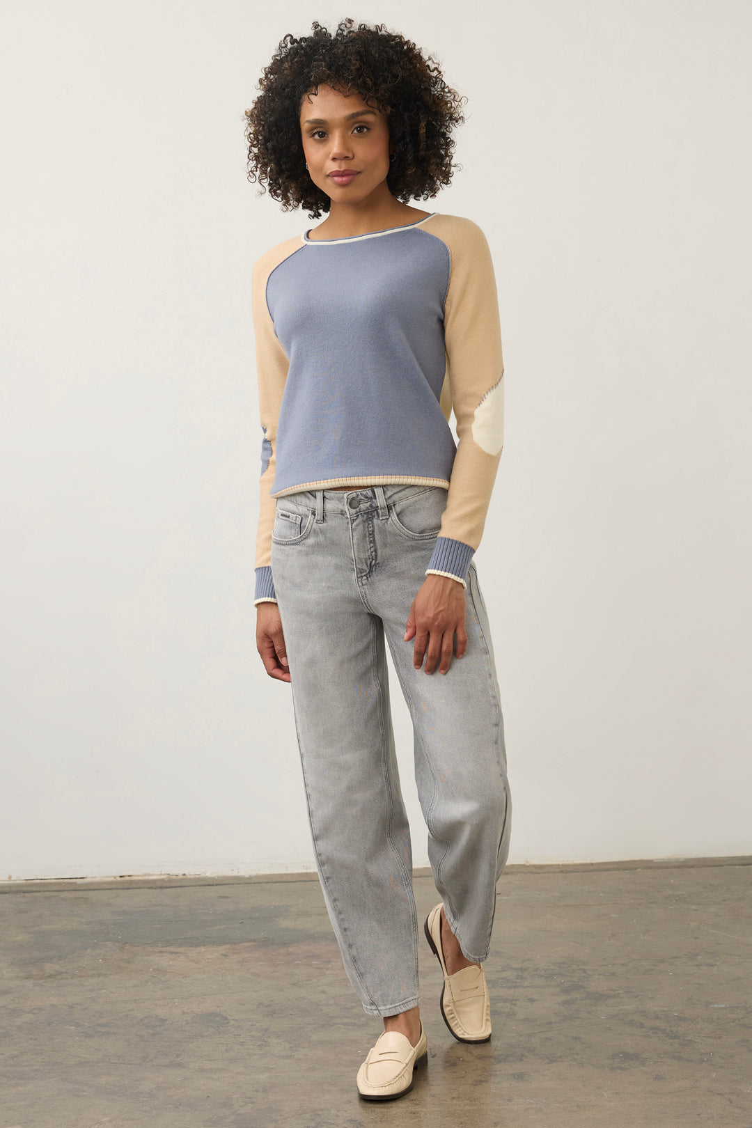A woman modeling a color-blocked knit sweater with a dusty blue body and beige sleeves, paired with light-wash jeans and cream loafers, standing against a plain light gray wall.
